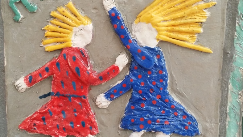 Clay mural of two blond girls in red and blue polka dot dresses dancing exuberantly, hair streaming out behind them.